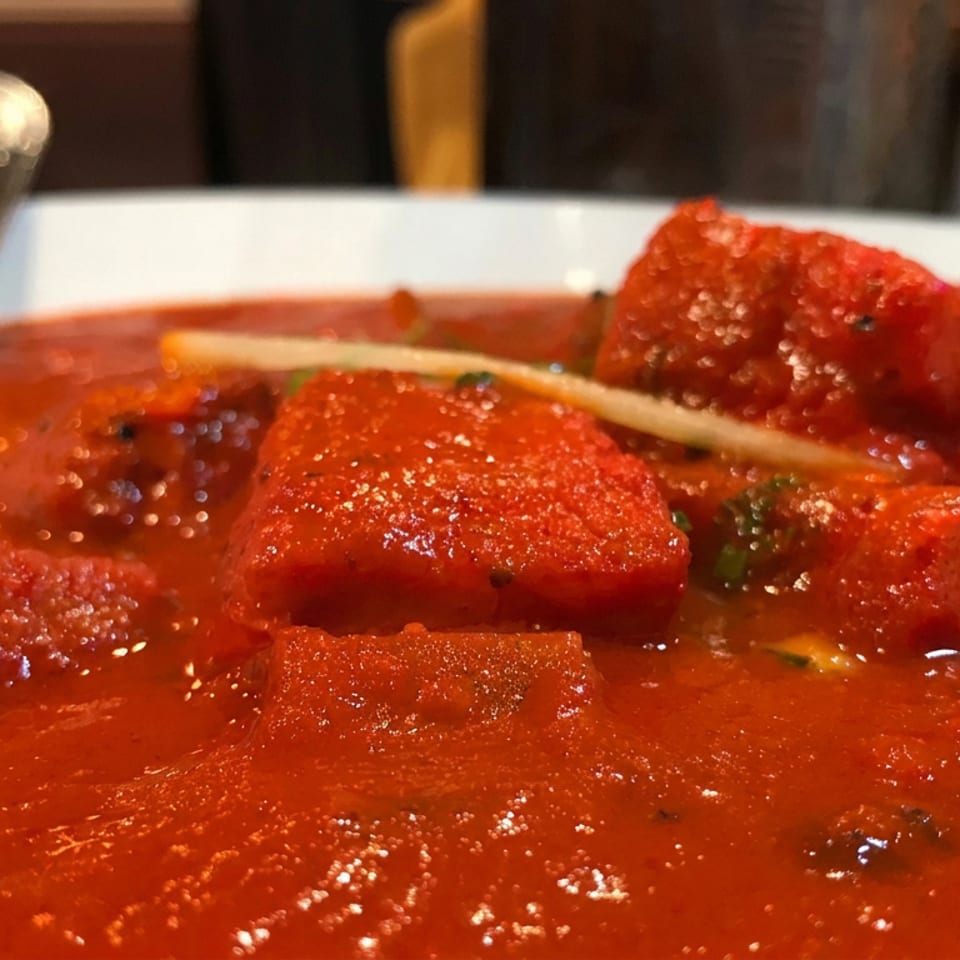 Close-up of red-coated paneer pieces simmering in a glossy tomato-based curry, garnished with herbs and served with a spoon.