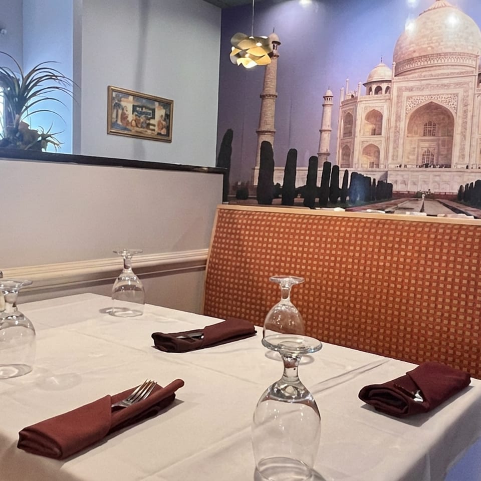 Elegantly set restaurant table with white linens, burgundy napkins, inverted wine glasses, and a Taj Mahal mural backdrop.