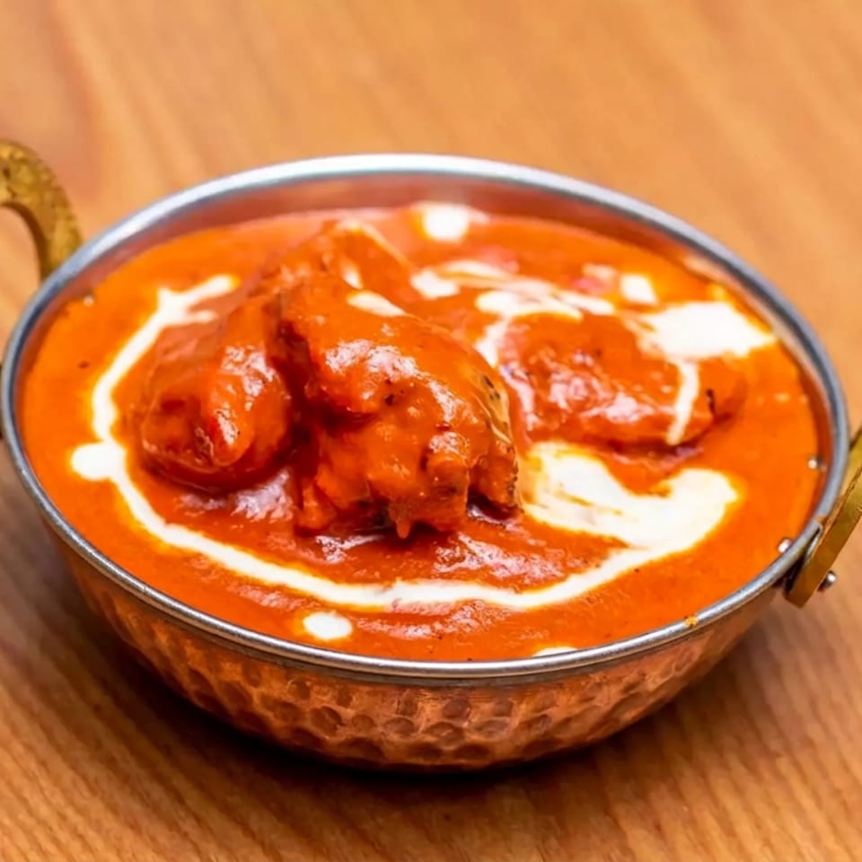 Indian butter chicken in creamy tomato sauce, garnished with cream, served in a brass-handled bowl on a wooden table.