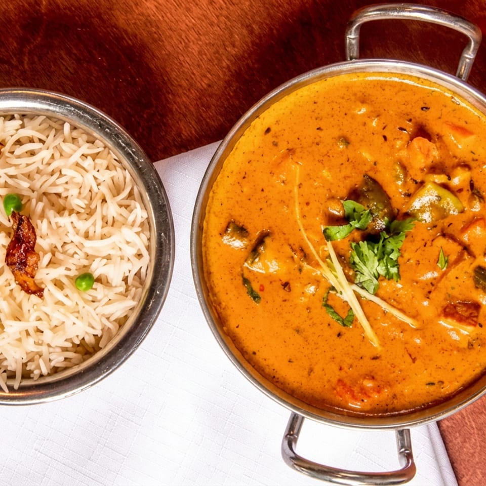 Overhead view of basmati rice with peas and fried onions beside creamy orange curry garnished with cilantro and ginger.